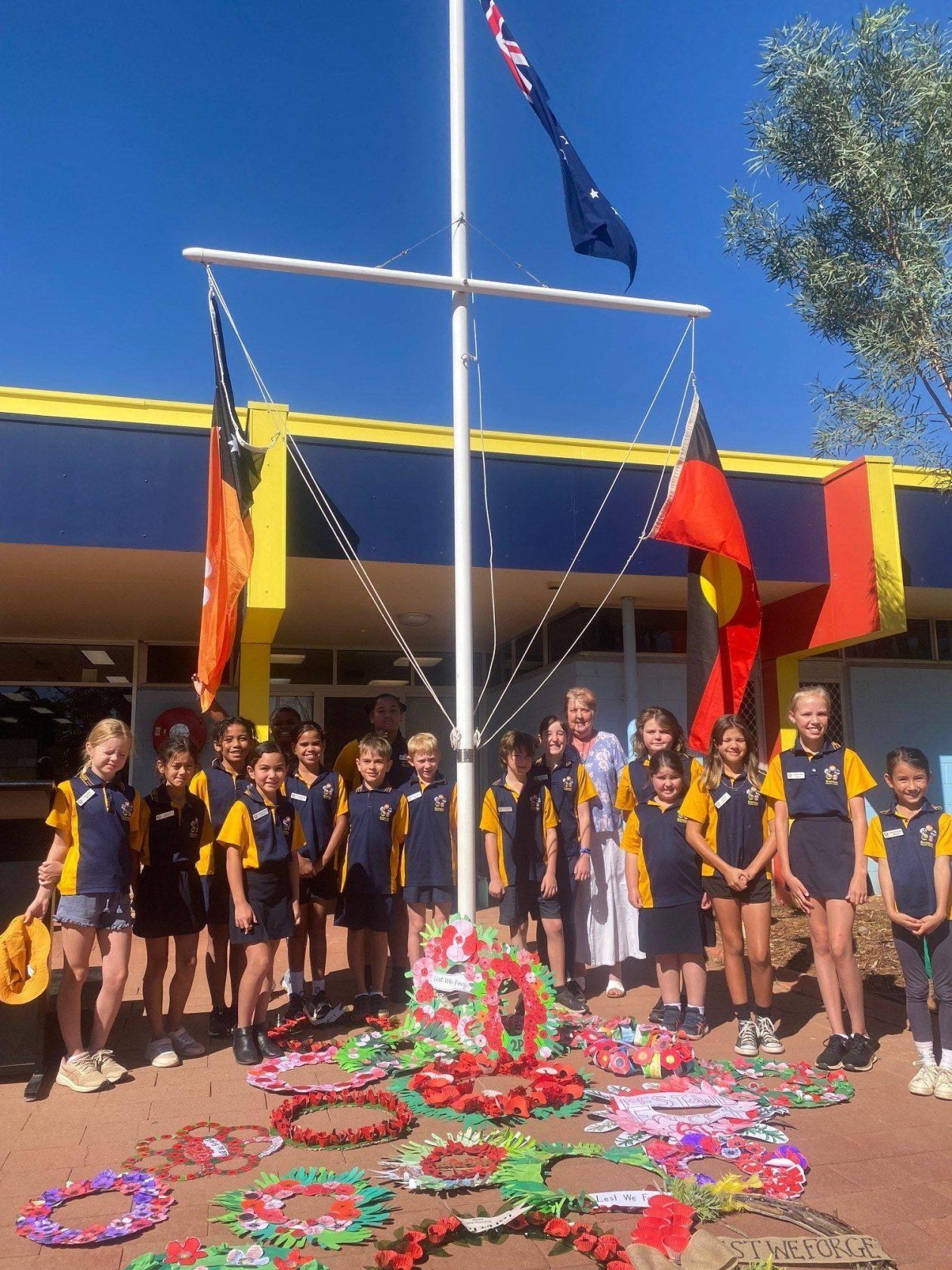 Schoolchildren stand by wreaths under a flagpole, with Australian and Aboriginal flags.