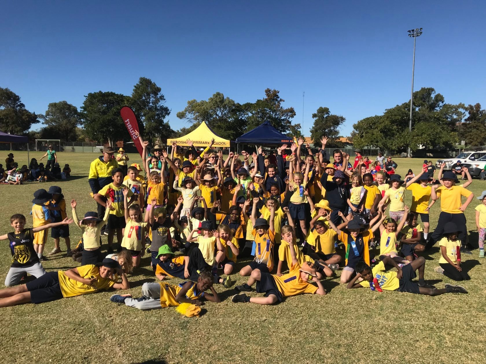 Large group of children in yellow shirts on a grass field, posing with smiles on a sunny day.