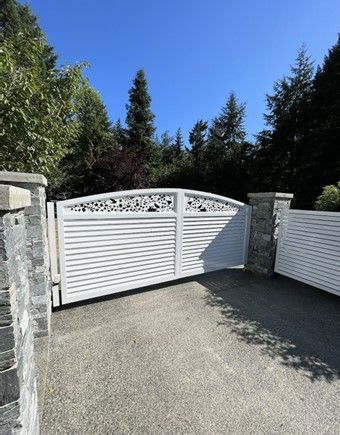 White driveway gate between stone pillars, set against trees and a blue sky.