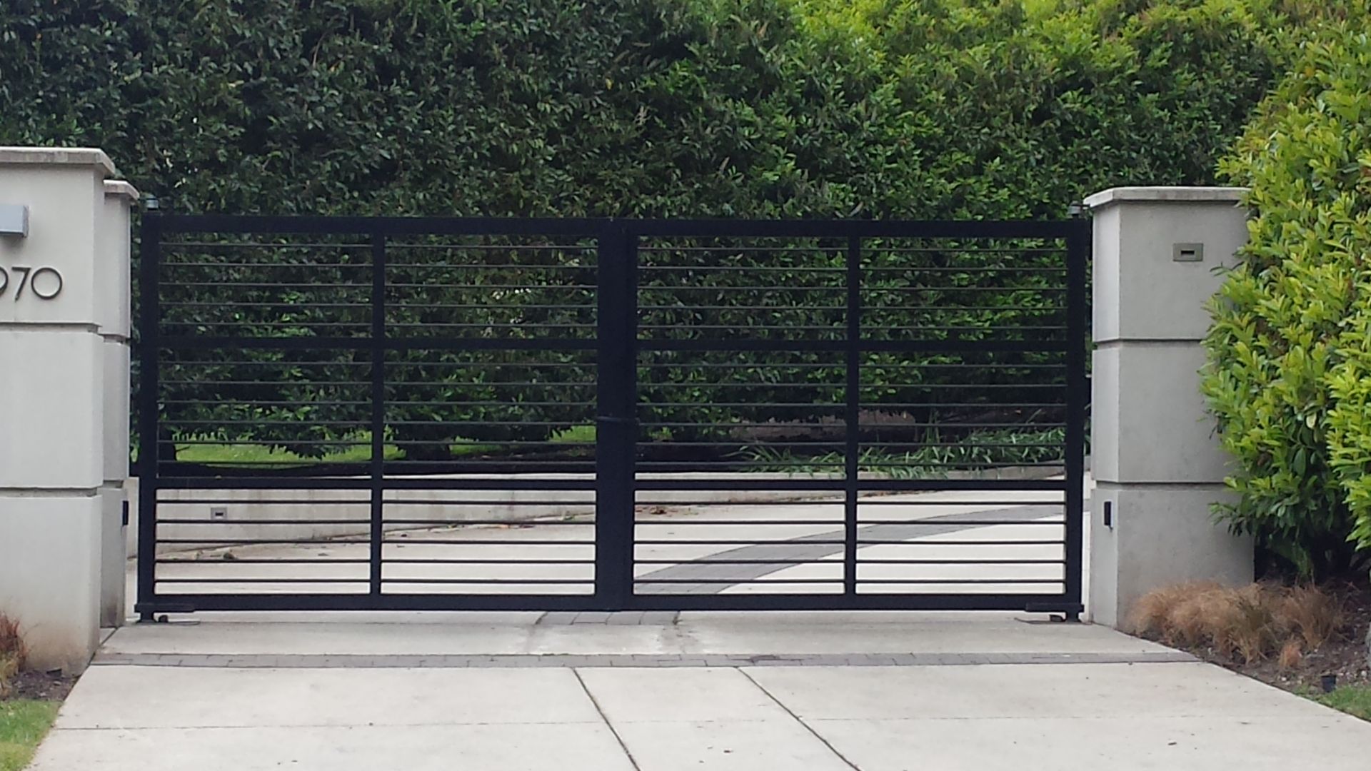Black metal driveway gate between stone pillars, with greenery in the background.