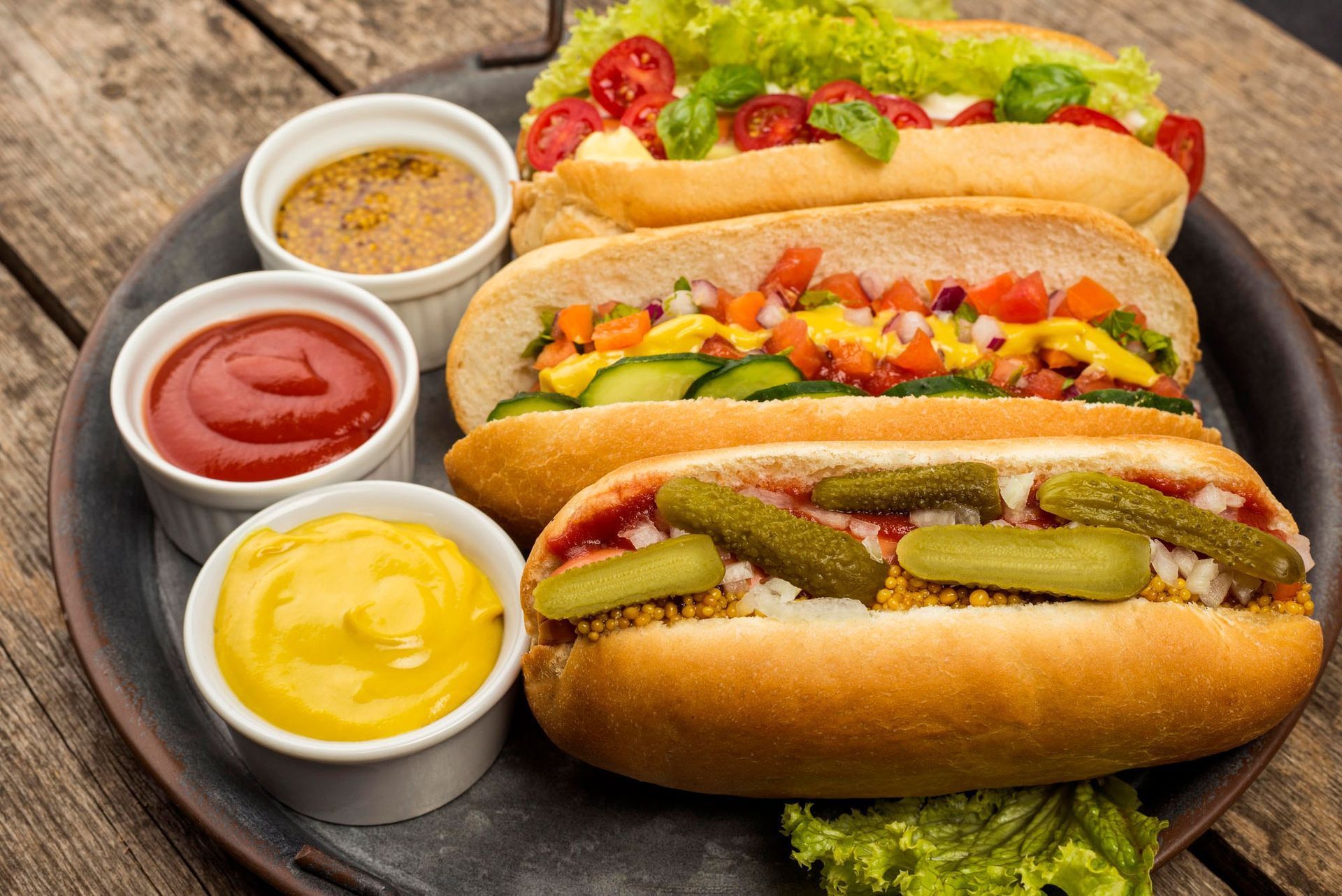Three hot dogs with vegetables and condiments on a plate on a wooden table.