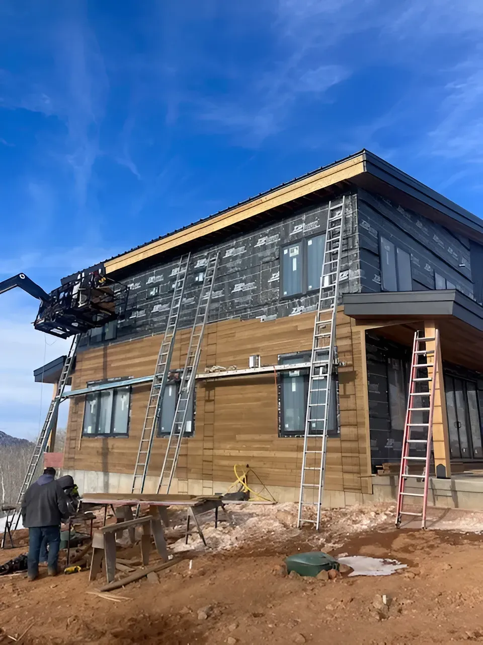 Two-story house under construction with workers, ladders, exposed insulation, and a blue sky backdrop
