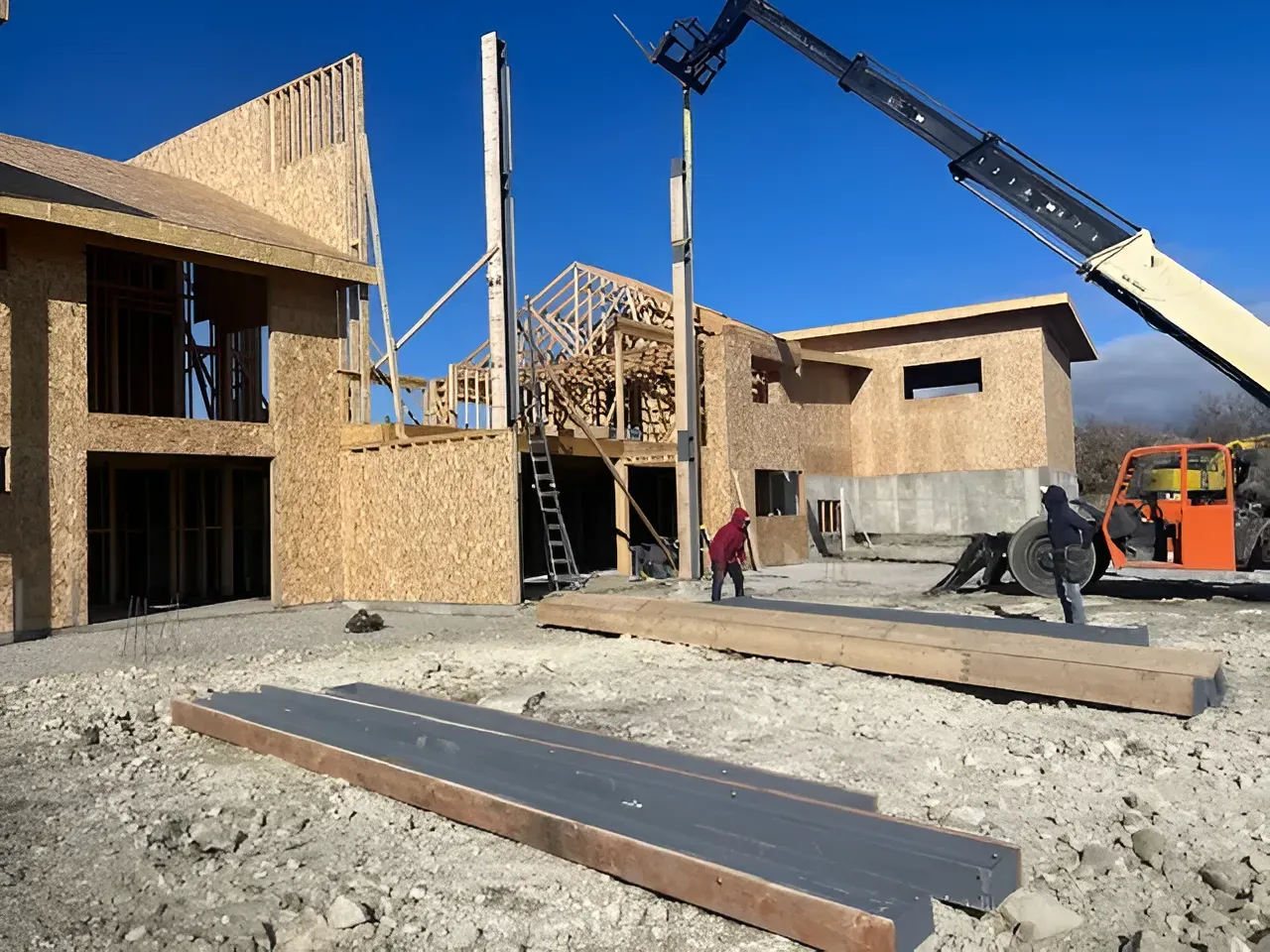 Wood-frame houses under construction with workers, crane, and building materials on a gravel lot