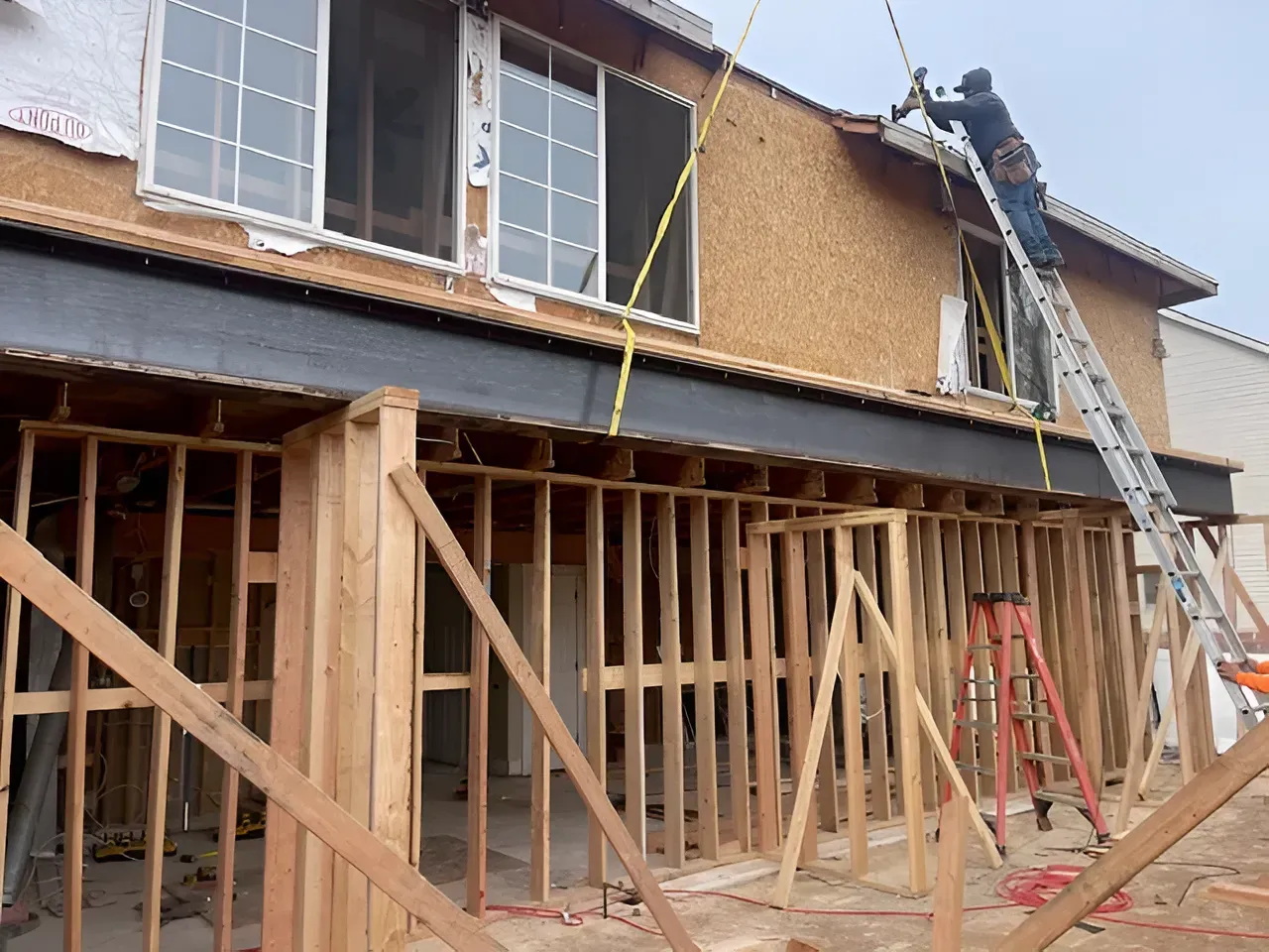 House under construction with exposed wooden framing and workers on ladders and scaffolding.