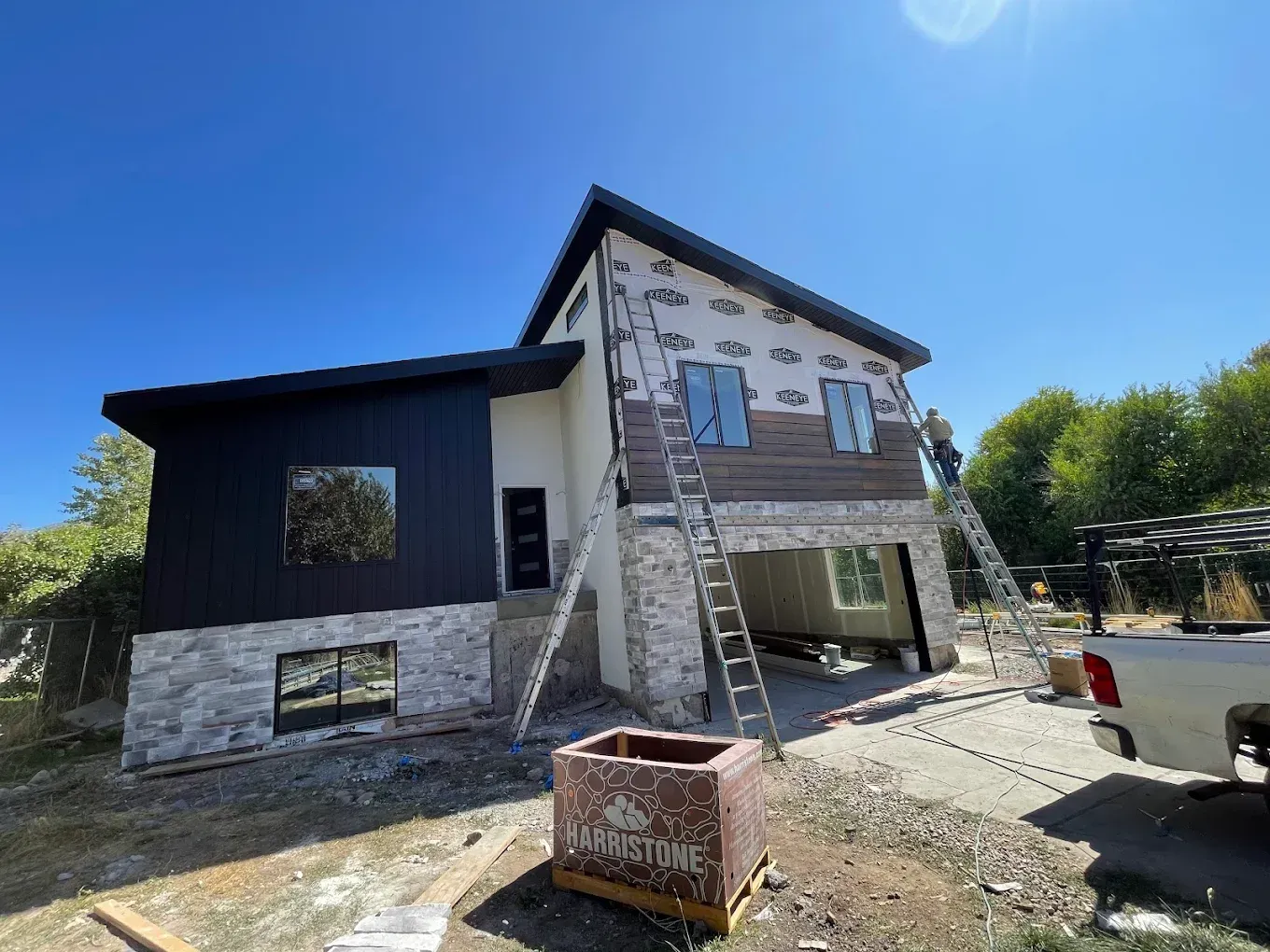 Two-story house under construction with stone siding, ladders, and a pickup truck in front under a blue sky