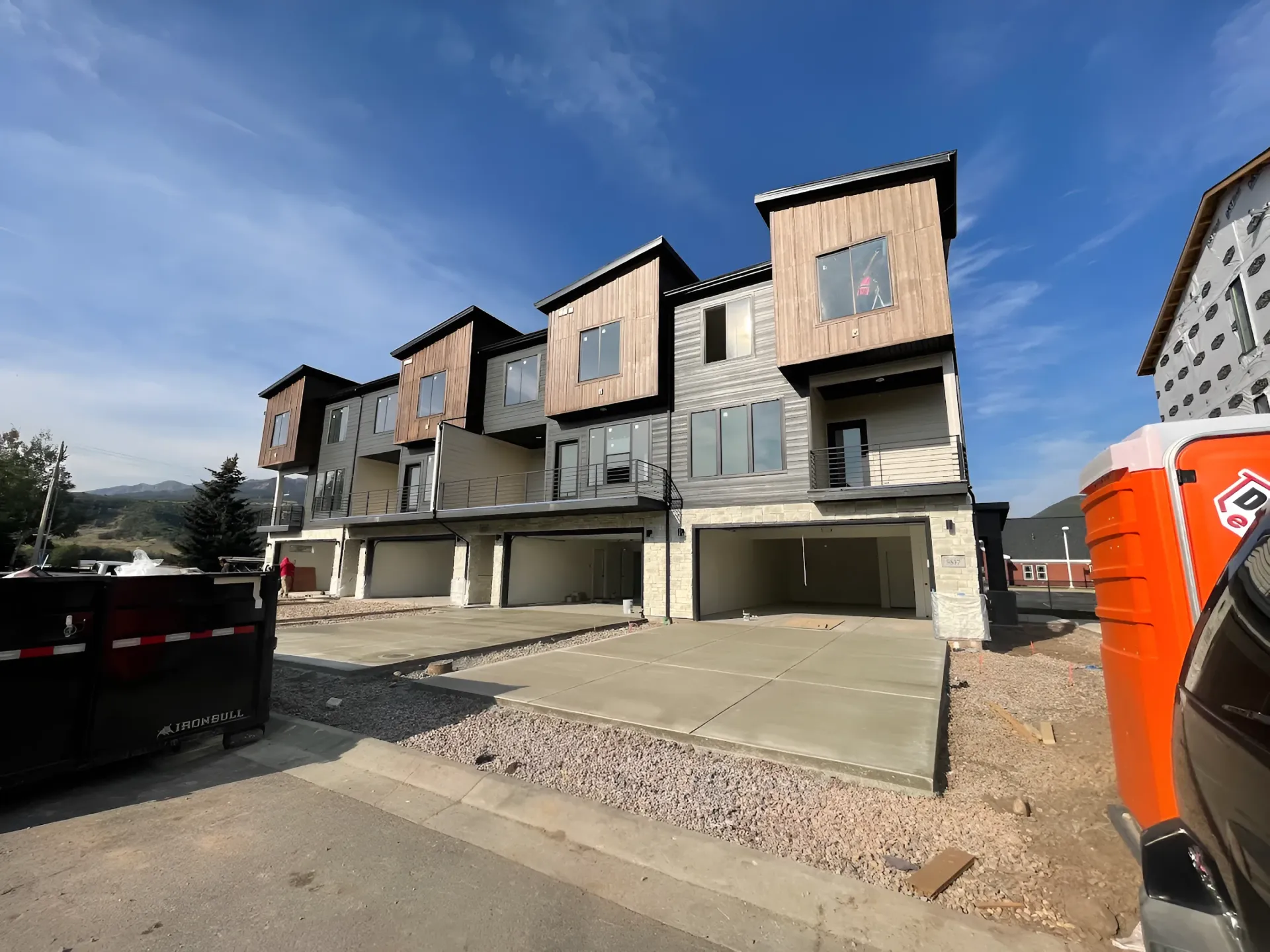 Row of unfinished modern townhomes with garages under a blue sky, viewed from a construction site