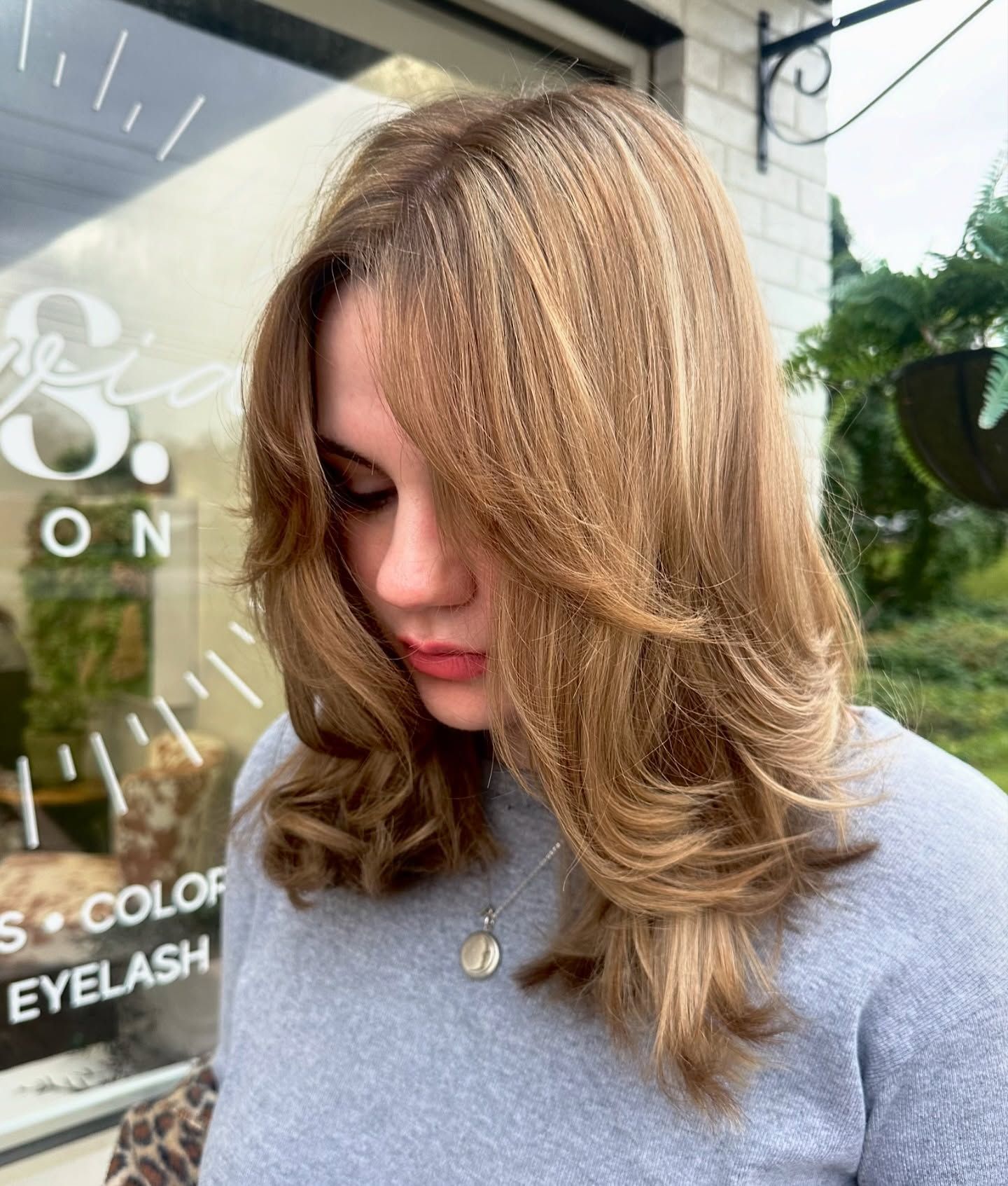 Woman with layered, highlighted brown hair looking down, standing near a shop window.