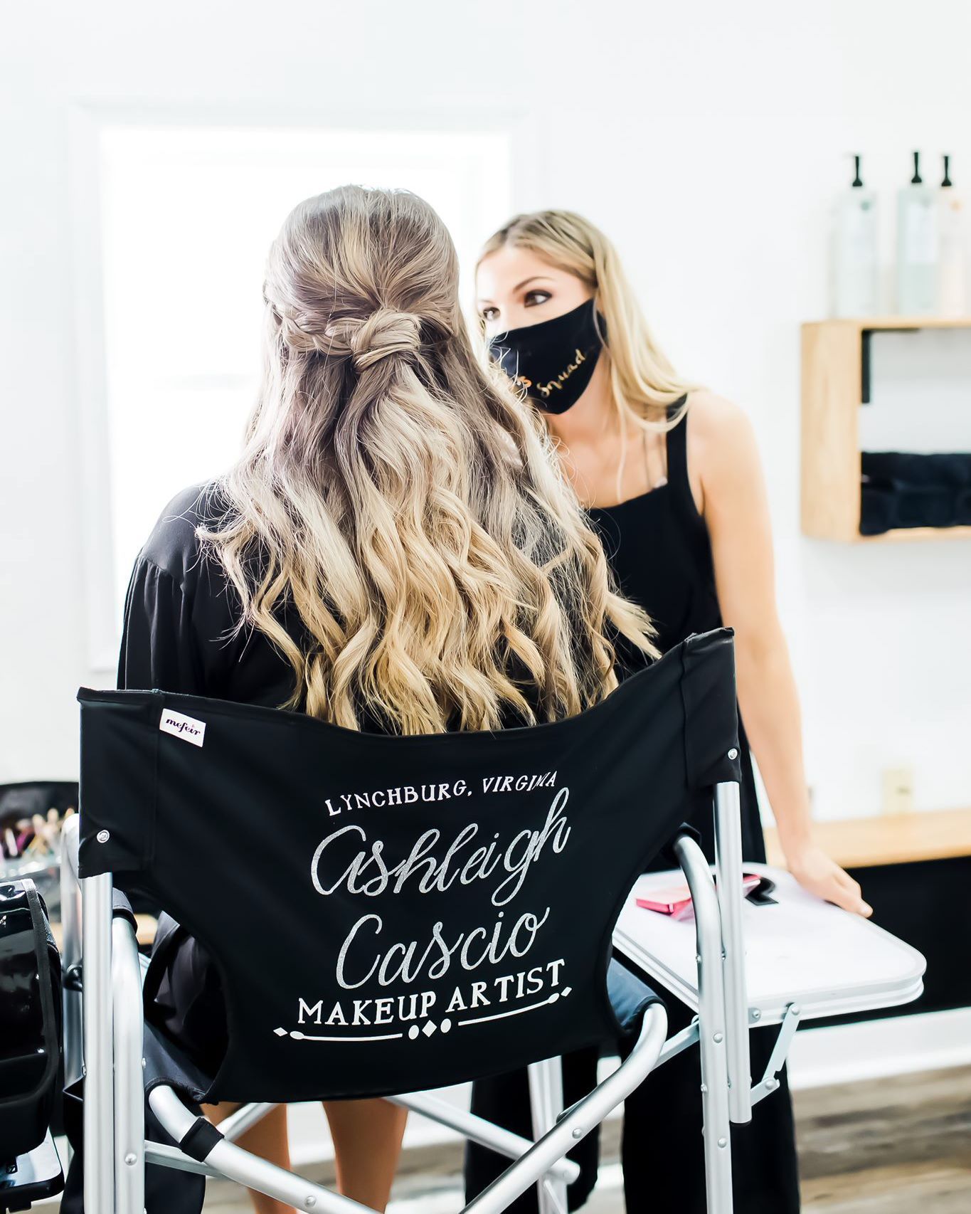 Makeup artist and client in a bright studio. Artist, in a black mask, stands over client seated in a director's chair.