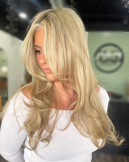 Woman with blonde crimped hair, wearing white off-the-shoulder top, indoors near a salon window.