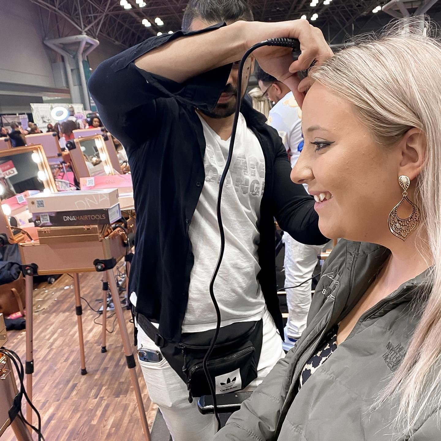 Makeup artist styling a smiling woman's hair in a brightly lit room. A makeup station is visible behind them.