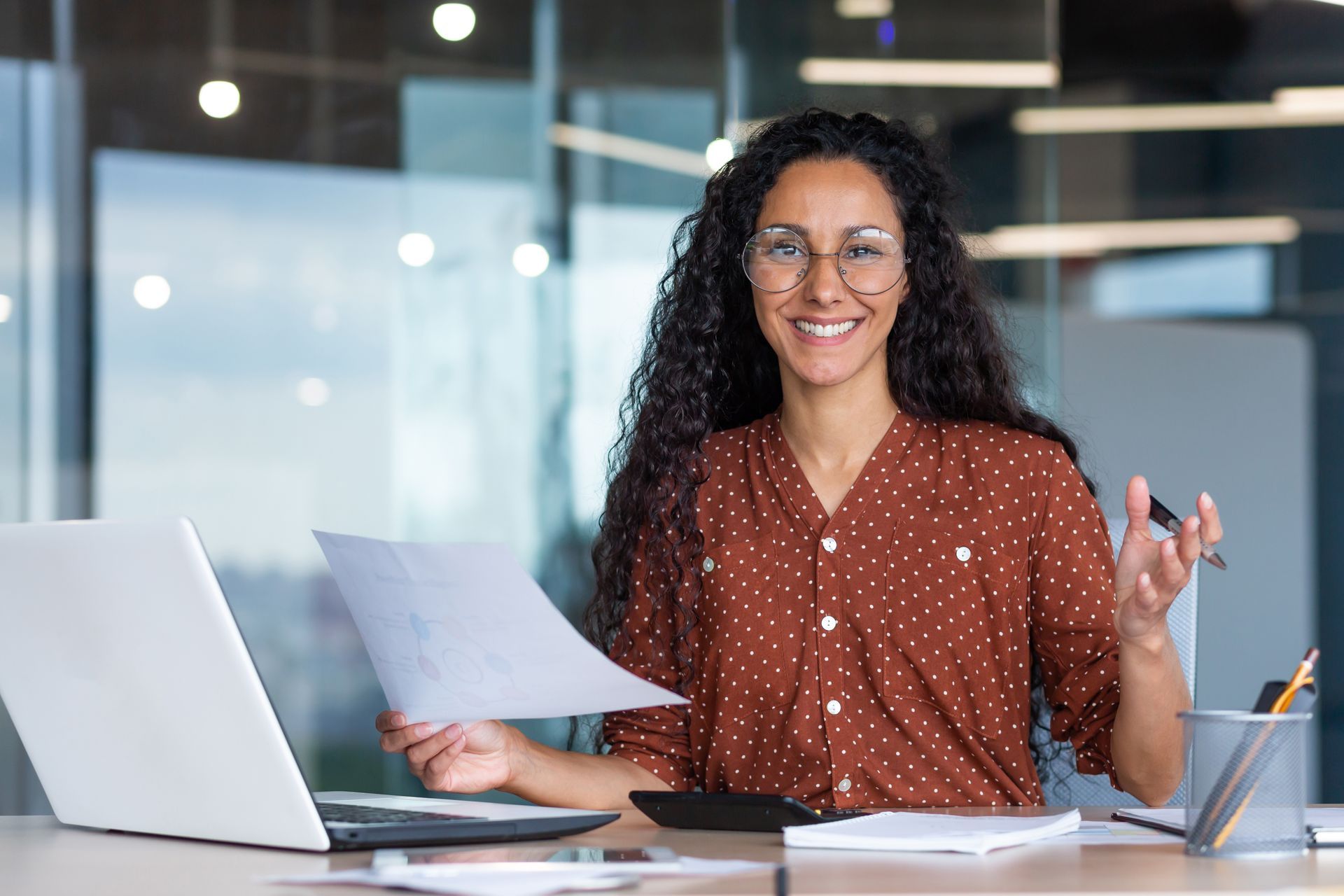 Woman in glasses smiling, holding papers, sitting at a desk with a laptop and calculator.