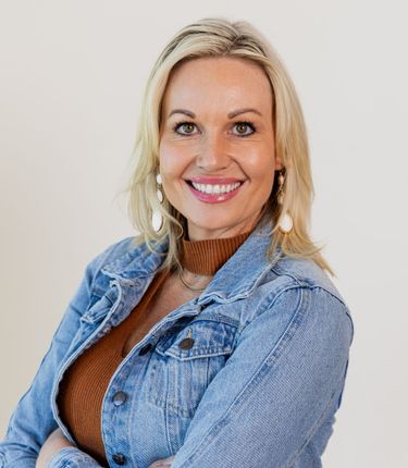 A woman wearing a denim jacket and a brown sweater is smiling for the camera.