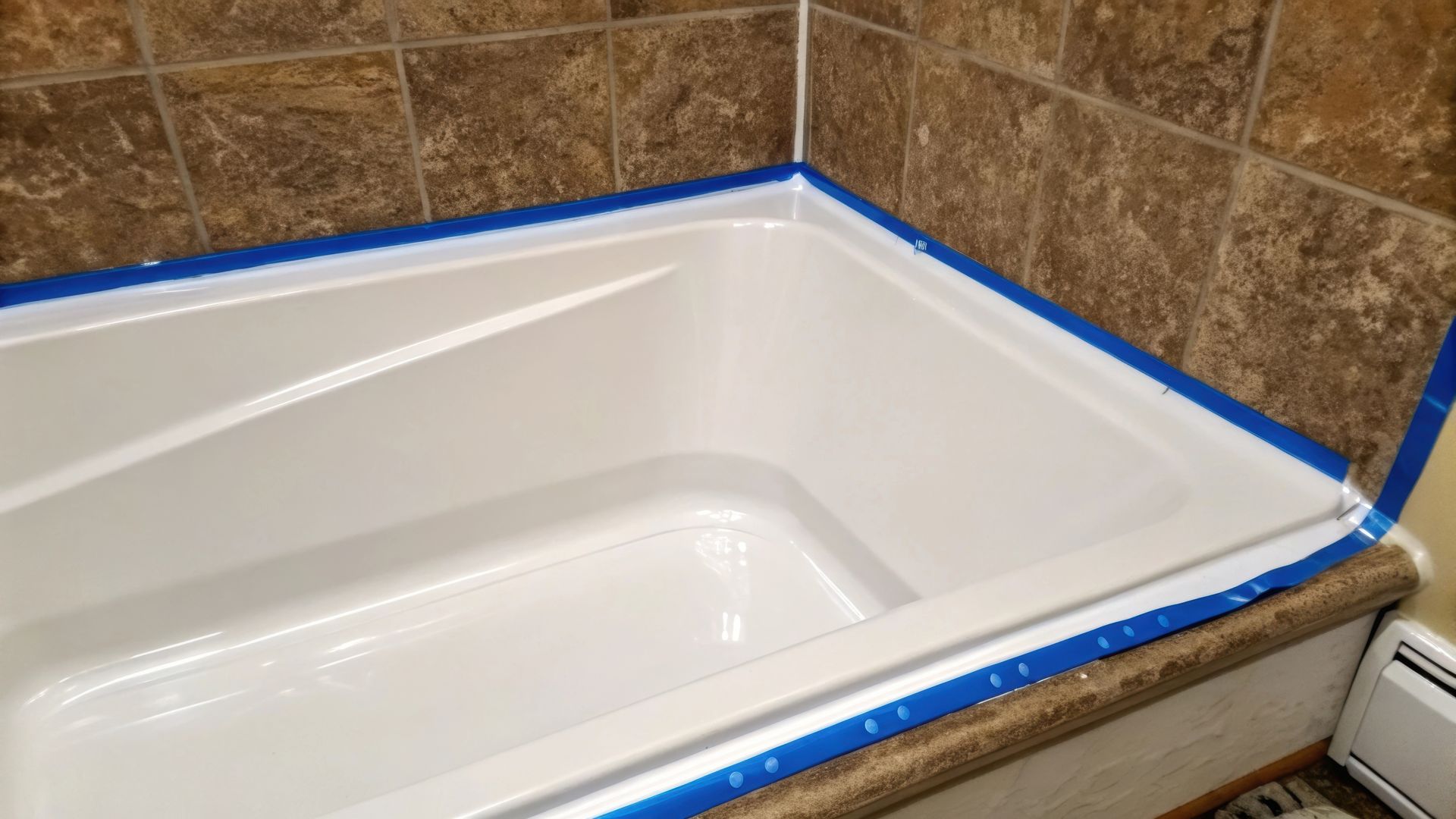 White bathtub with blue painter's tape along the edges, ready for caulking against brown tile walls.