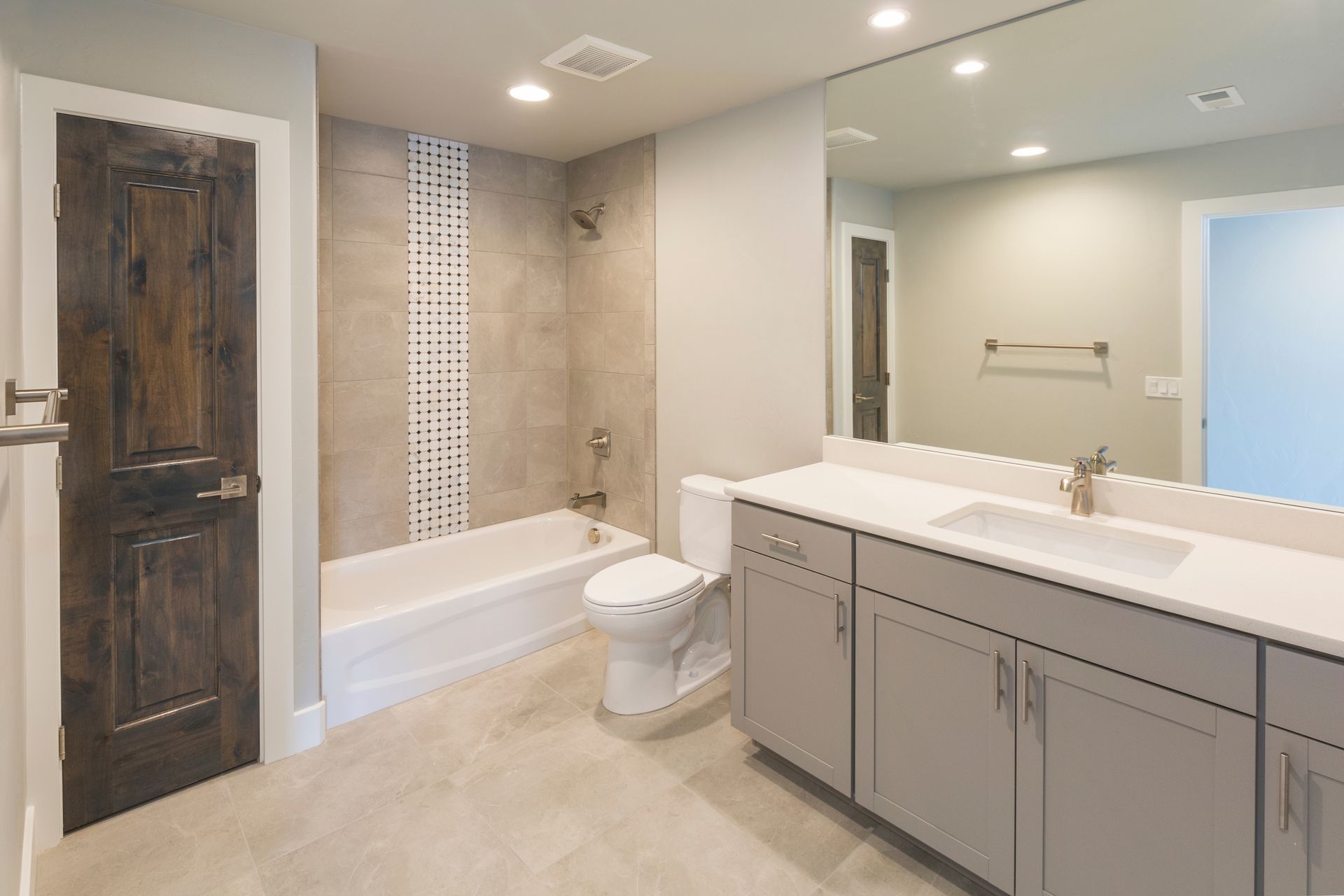 Bathroom with a gray vanity, white countertop, toilet, bathtub, and dark wooden door.