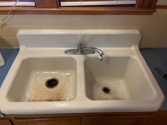 White double sink with rust stains, chrome faucet, and blue countertop.  Cabinet below, window in the background.