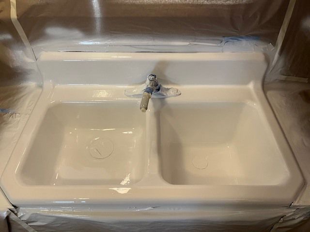 A white double-basin kitchen sink with a chrome faucet, masked with plastic sheeting for painting.