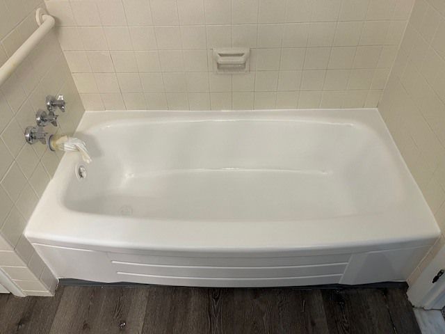White bathtub in a bathroom with a tiled wall and chrome fixtures. The floor is a dark wood-look laminate.