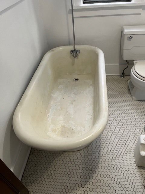 A slightly dirty, off-white bathtub in a bathroom with a shower head, toilet, and white tiled floor.