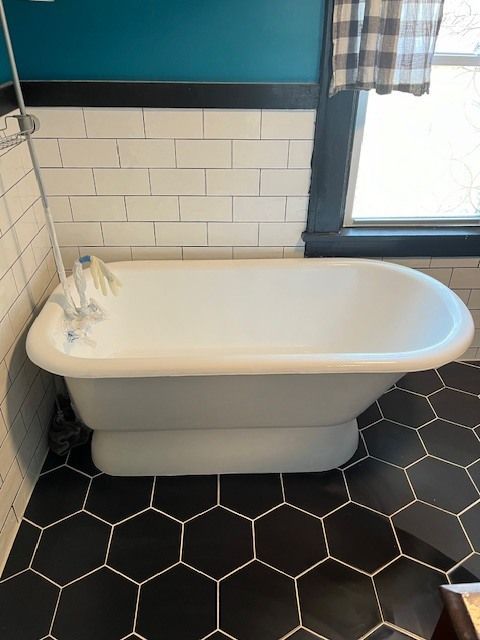 A vintage-style white bathtub sits in a bathroom with black hexagon tile flooring and white subway tile walls.