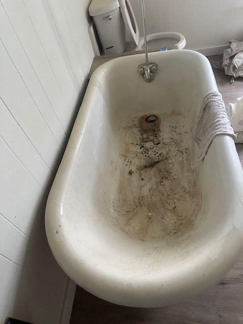 Dirty, vintage-style white bathtub with dark stains. A trash can and toilet are visible in the background near a white wall.