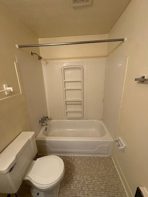Small bathroom with a white bathtub, toilet, and shower with a built-in shelf. Beige walls and a tile floor.