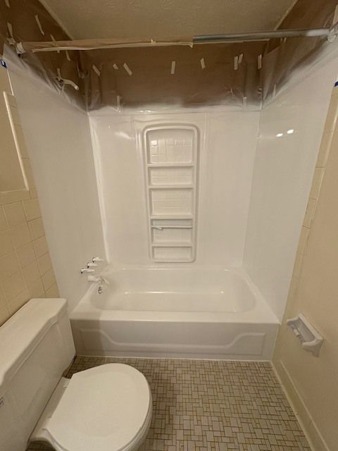 Bathroom with white tub and shower surround, toilet, and beige walls. The floor has small square tiles.