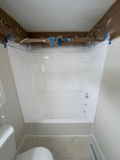 White bathroom shower stall with installed fixtures, viewed from the toilet; protective plastic attached to the ceiling.