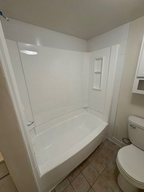 White bathtub and shower unit with a built-in shelf, beside a white toilet, in a bathroom with tiled floor.