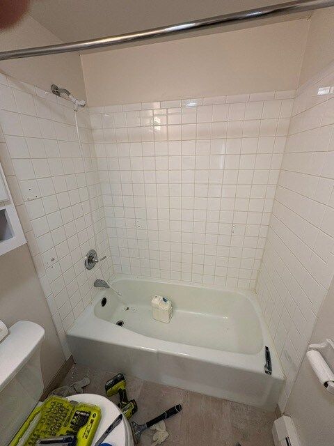 White tiled bathroom with a bathtub and showerhead, under a shower rod. Tools and toilet are visible on the floor.