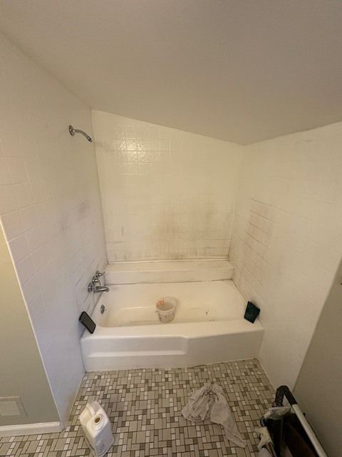 A dirty white bathroom with a tub and shower. Reddish stains are visible on the walls, and the floor is tiled.