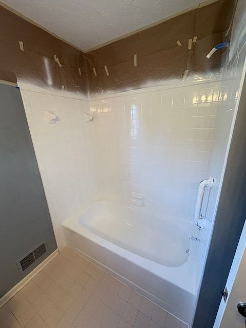 Bathroom with a white bathtub and shower surround, beige floor tiles, and partially painted walls with masking tape.