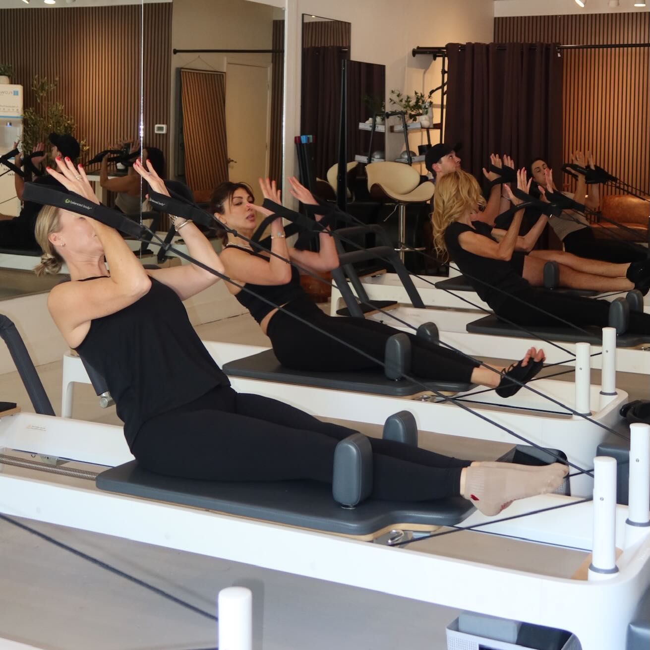 People exercising on reformer Pilates machines in a bright studio, wearing black workout clothes.