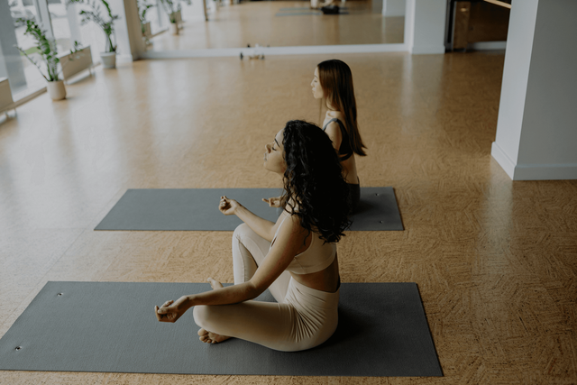 Two people seated cross-legged on yoga mats in a bright studio, facing forward.