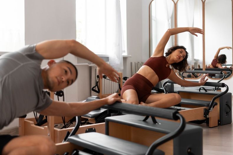 Two people stretching on Pilates reformers in a bright studio, one leaning toward the camera.