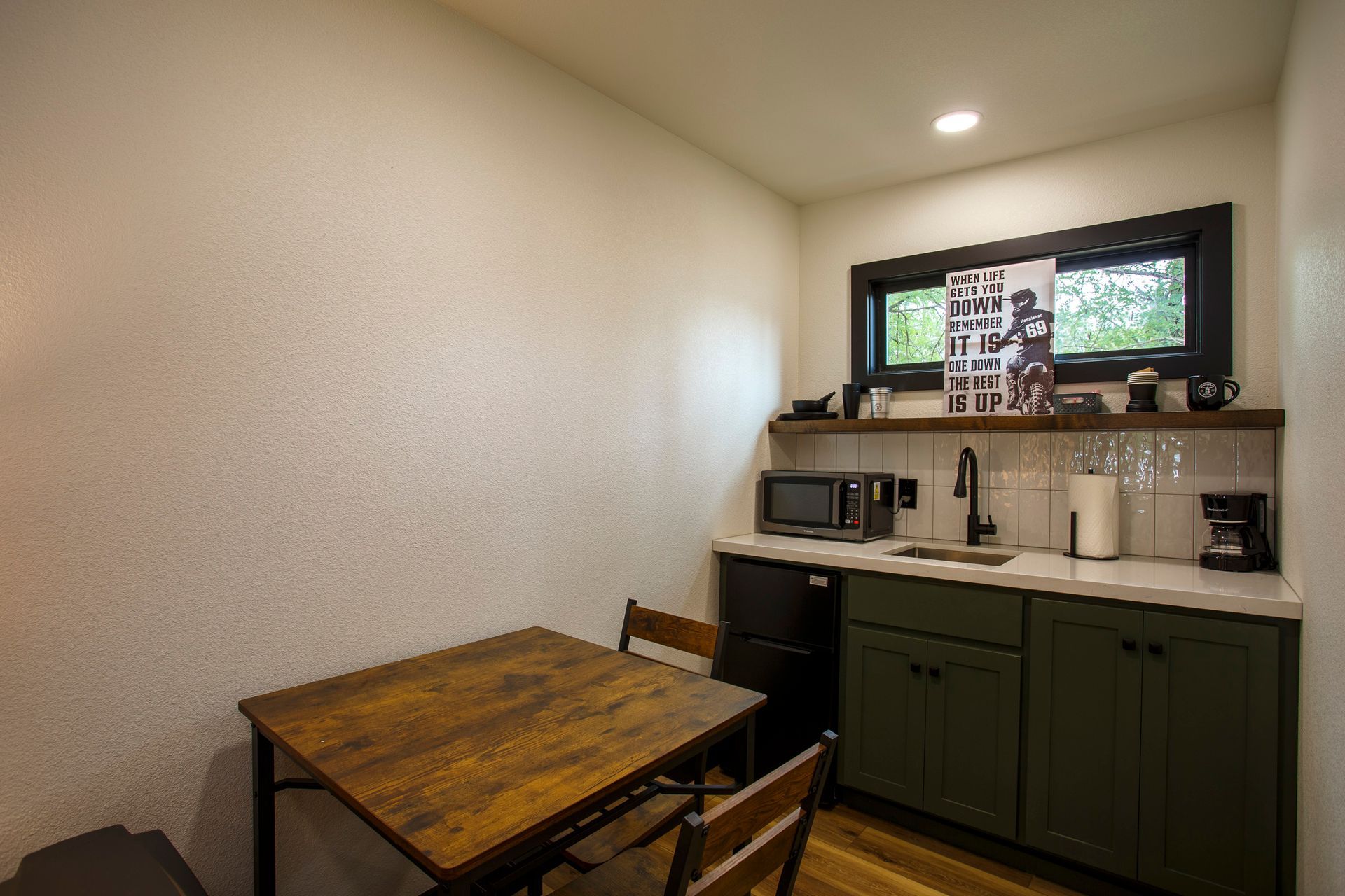 Small kitchen nook with a wooden table, two chairs, green cabinets, and a window above the sink.