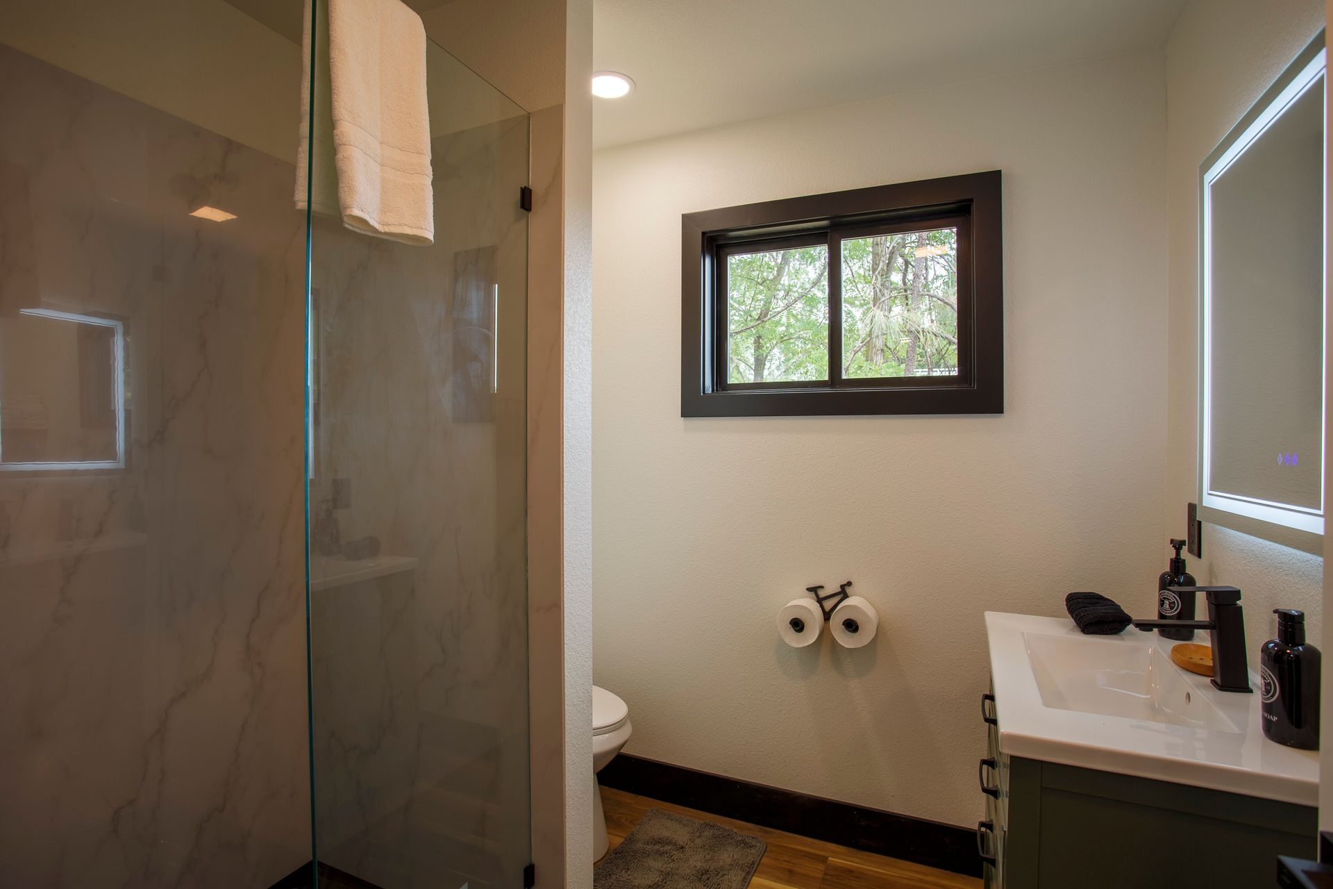 Small bathroom with glass shower, window, toilet paper holder, and vanity sink near a mirror.