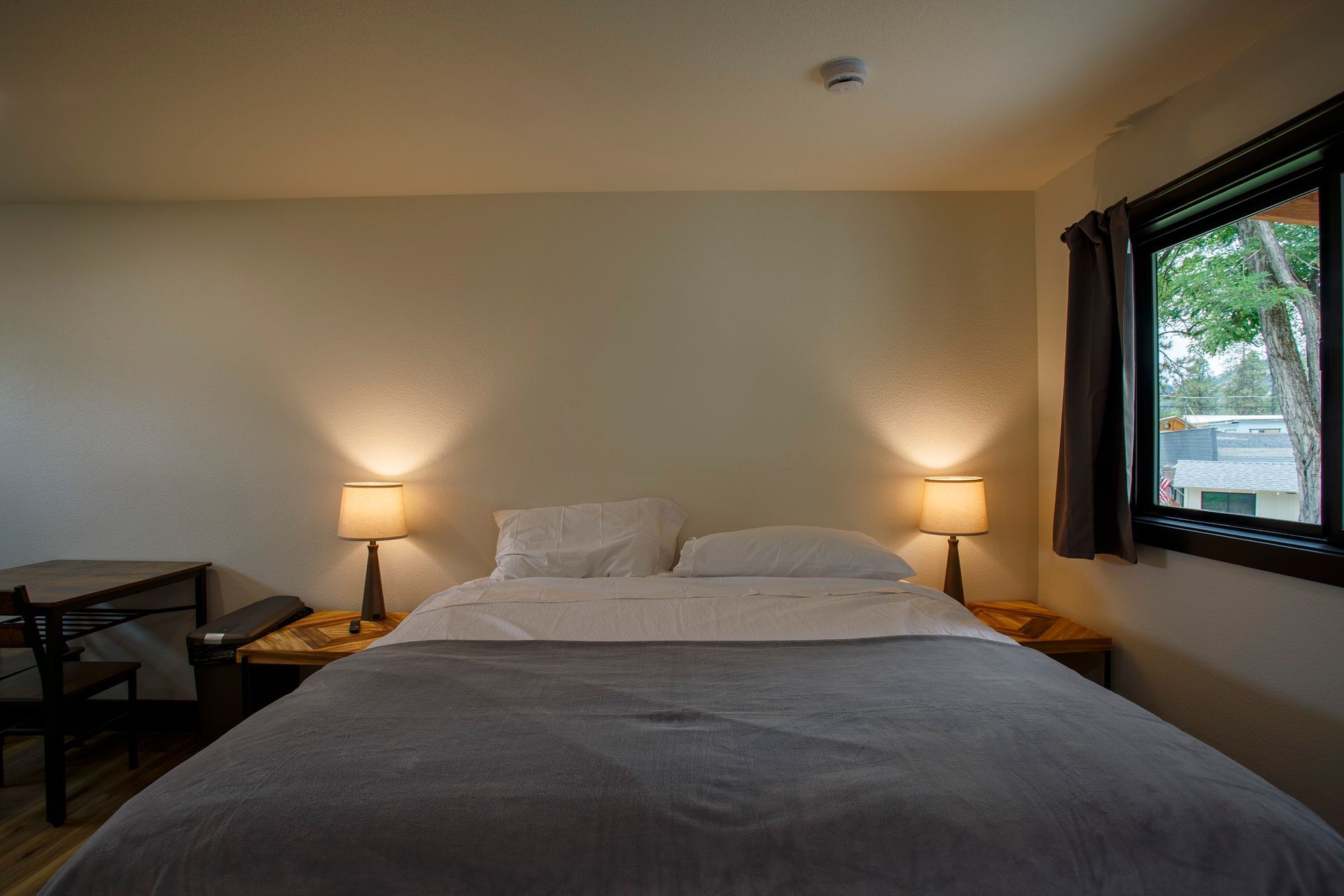 Hotel room with a neatly made bed, two bedside lamps, and a window showing greenery outside