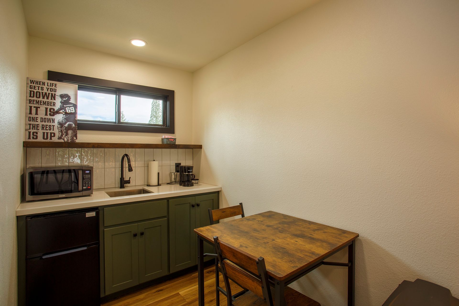 Small kitchen with sink, microwave, cabinets, window, and a wooden table against a white wall.
