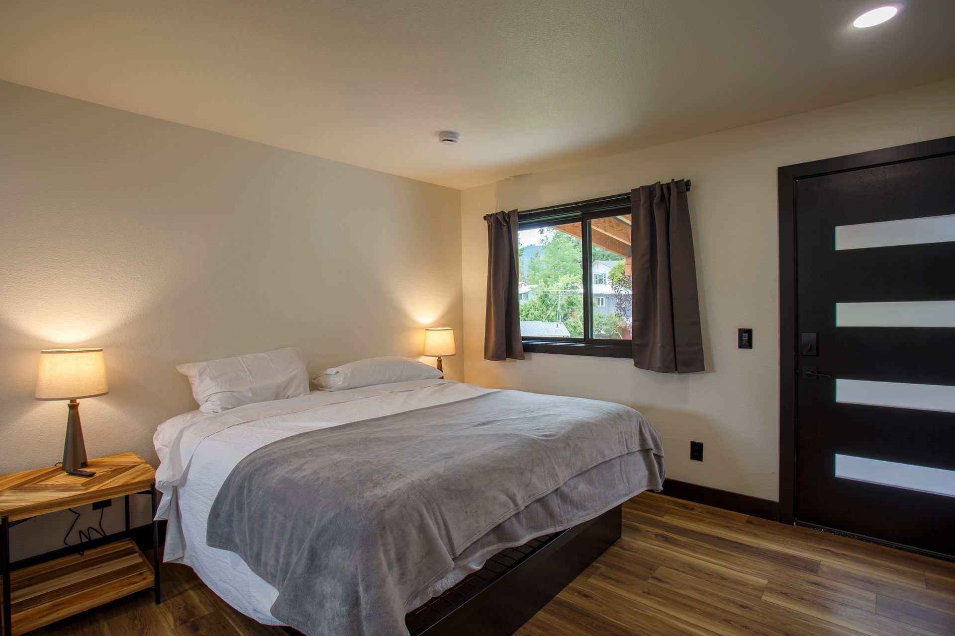Modern bedroom with a made bed, two lamps, window, and dark wood door
