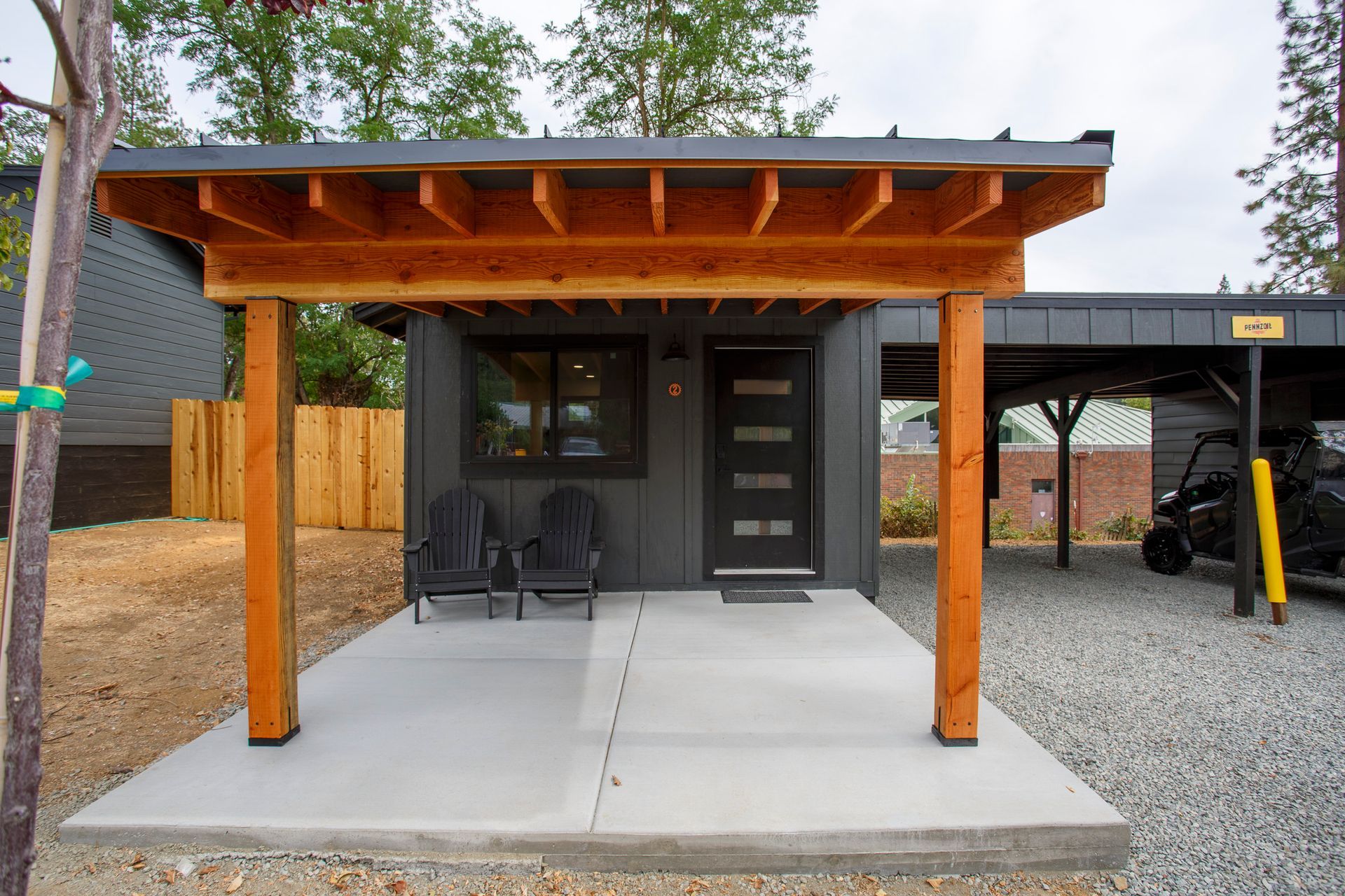 Small covered porch with wood posts, concrete slab, chairs, and a black door beside a gravel driveway.