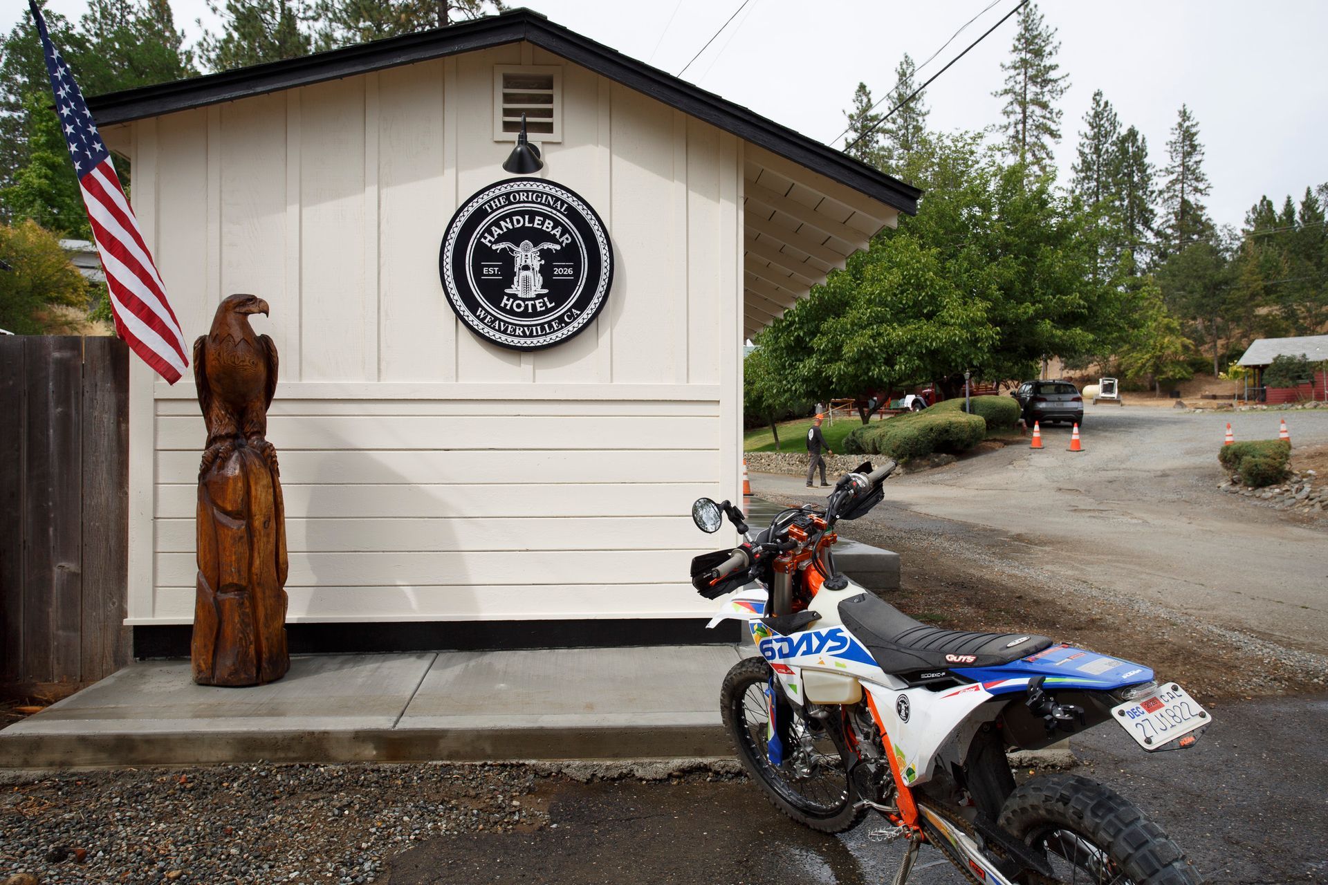 Motorcycle parked beside a building with a seal, American flag, and carved wooden statue in a forest setting