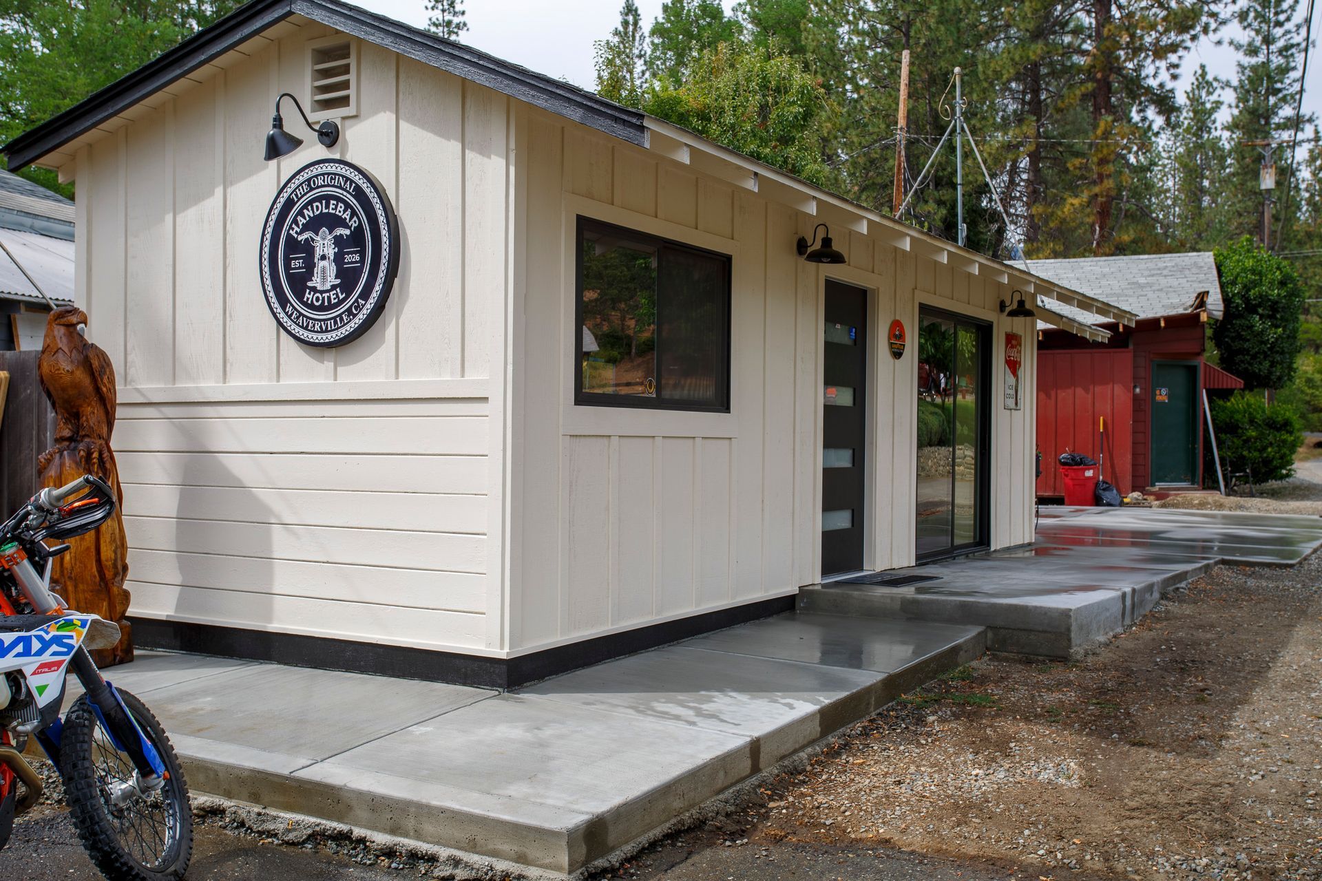 Small cream building with a circular sign, concrete steps, and a motorcycle parked outside in a wooded area