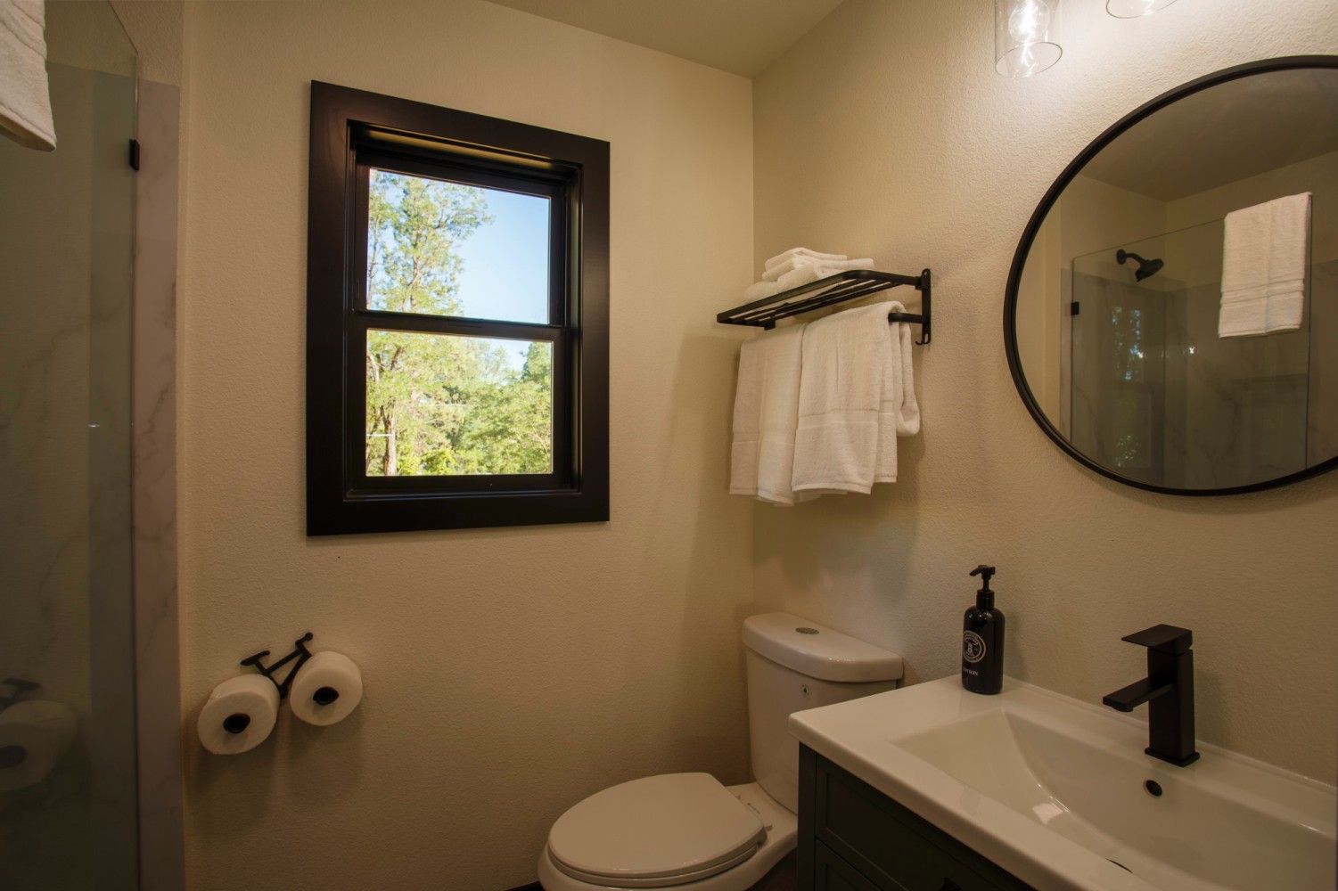 Small beige bathroom with window, toilet, vanity, round mirror, and towel rack