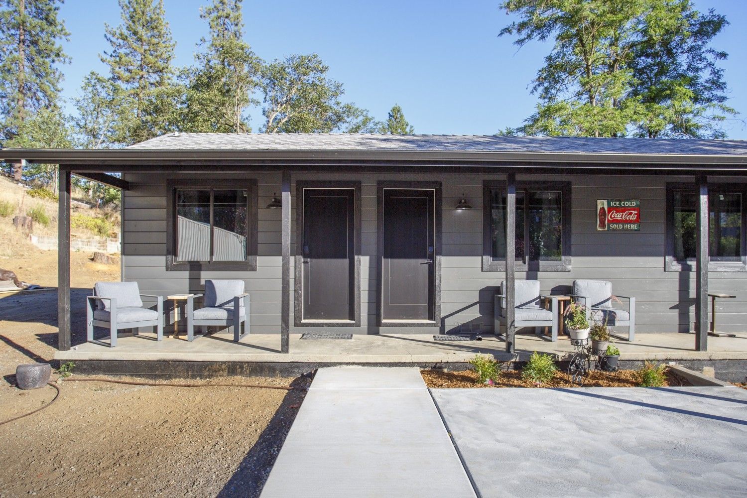 Small gray house with a front porch, patio chairs, and a concrete walkway in a sunny yard.