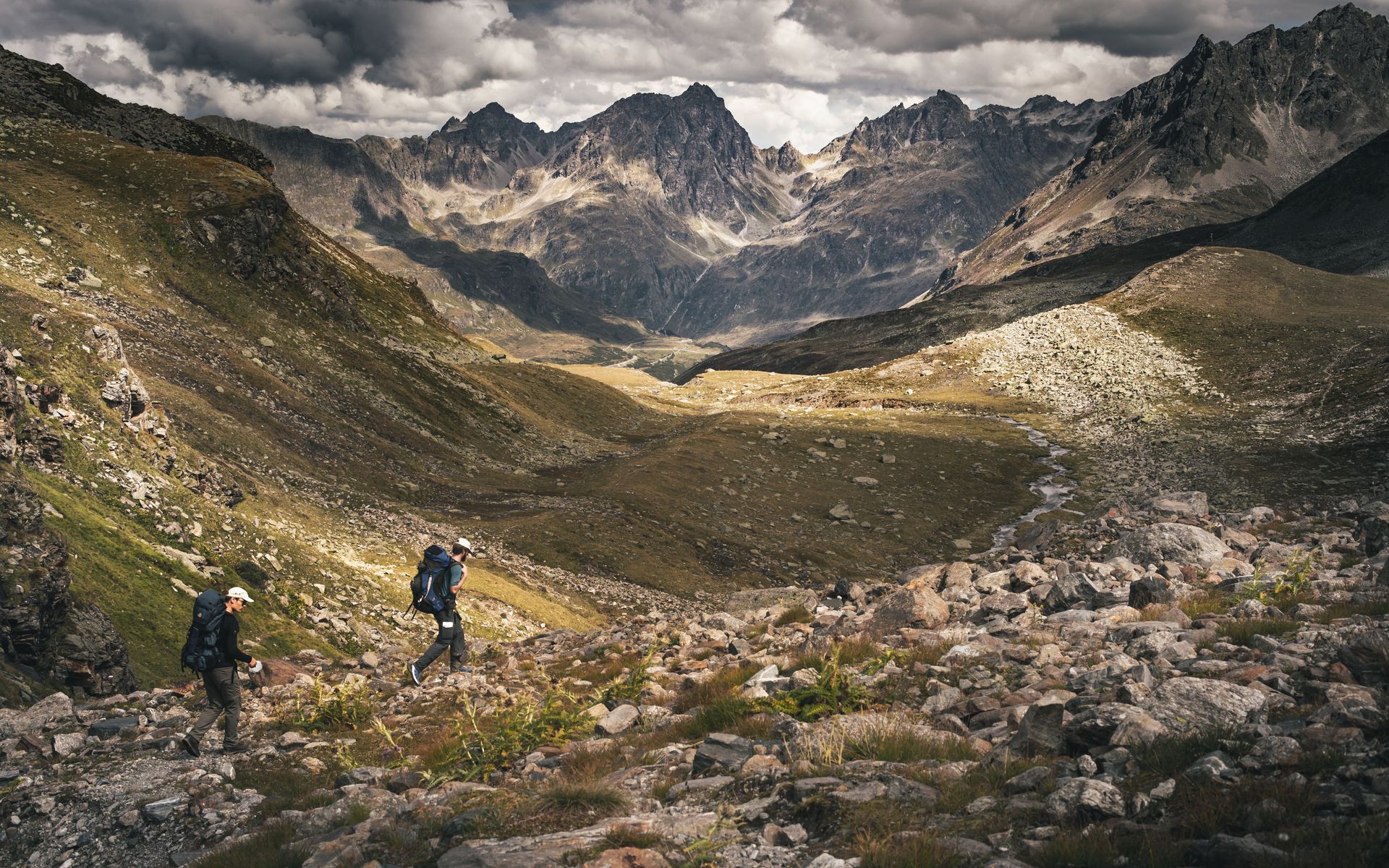 Hikers crossing a rocky mountain valley under dramatic cloudy skies