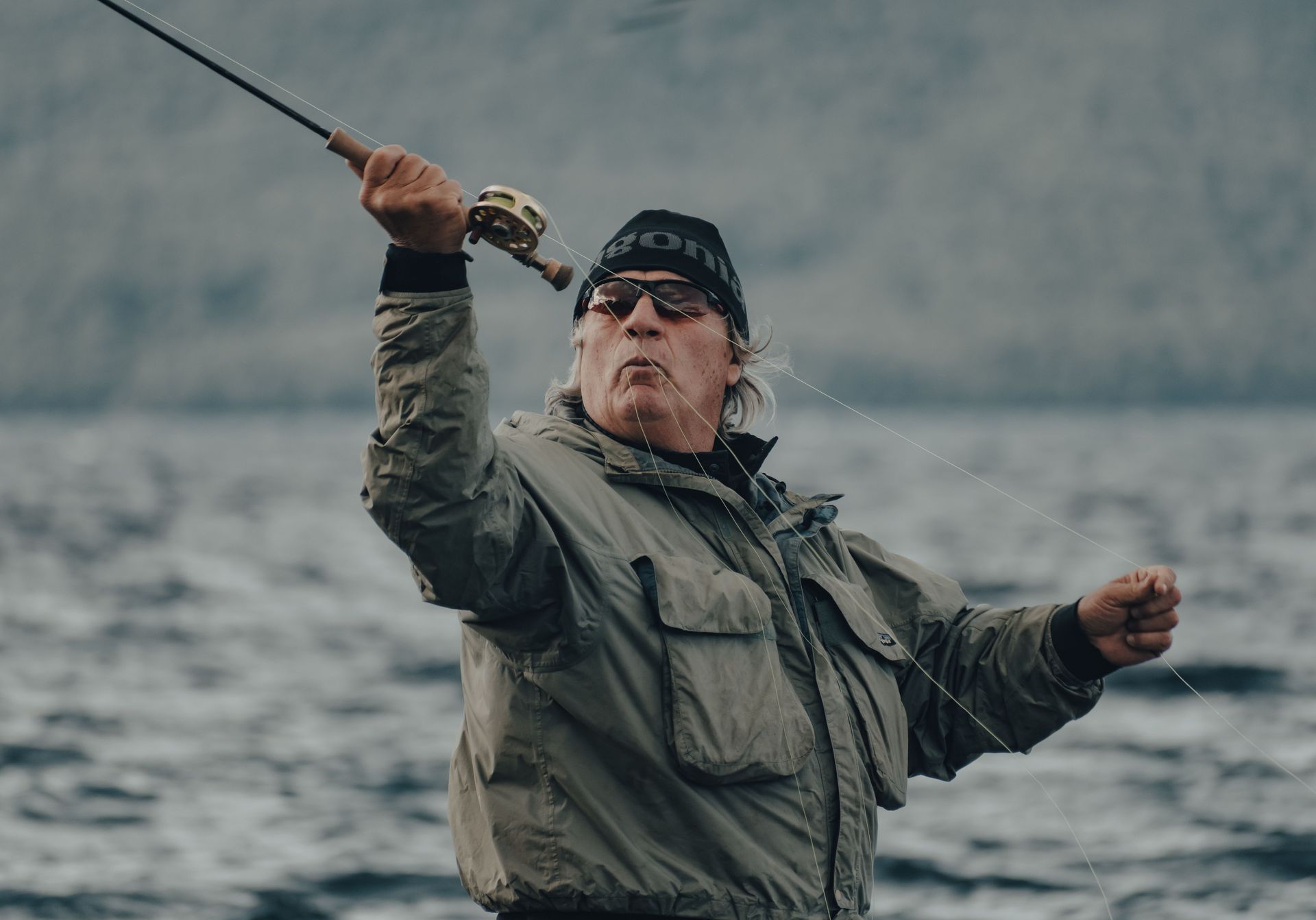 Person in a beanie and rain jacket holding up a fish by a misty lakeshore