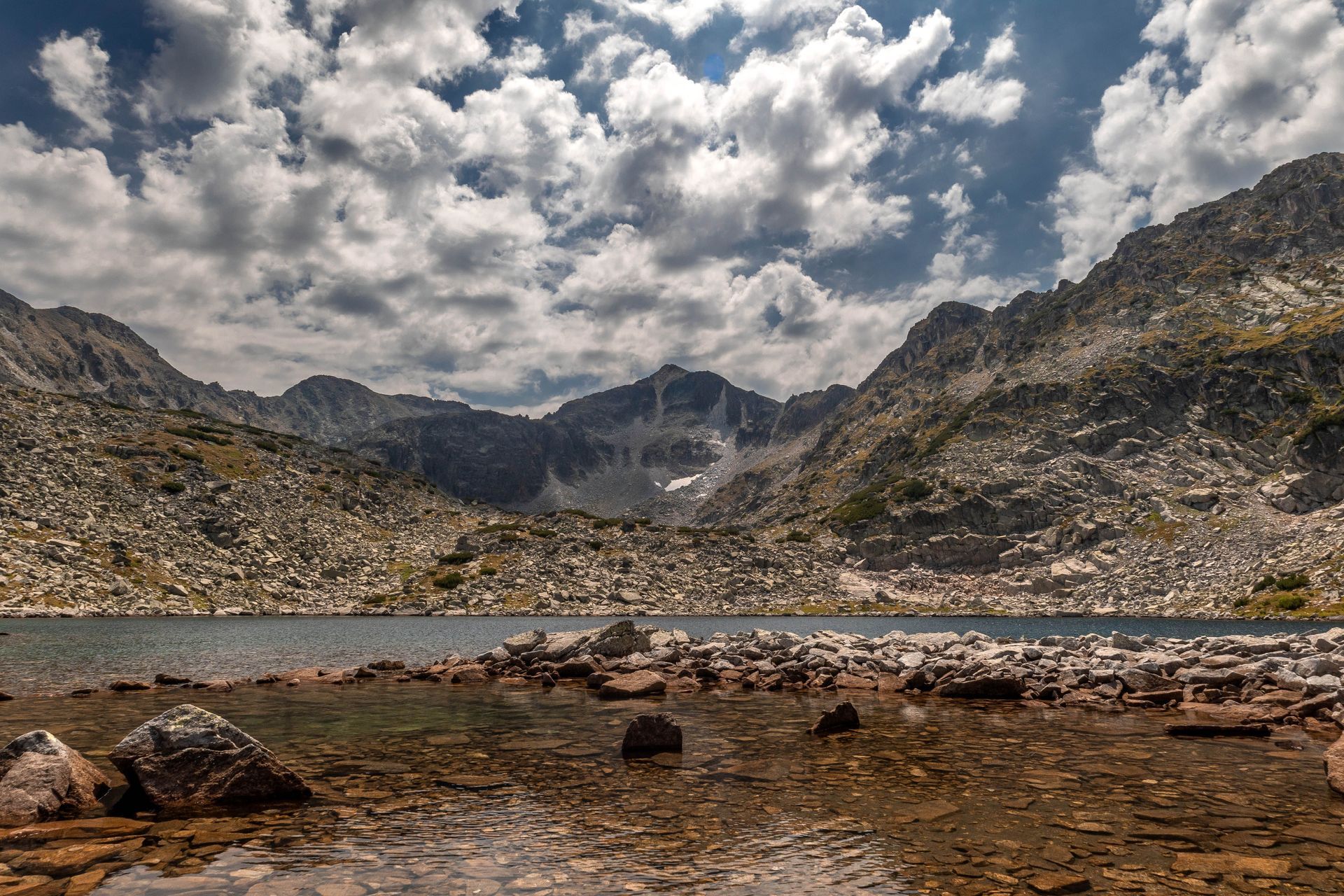 Rocky mountain lake beneath cloudy sky, with rugged peaks and shallow water in the foreground