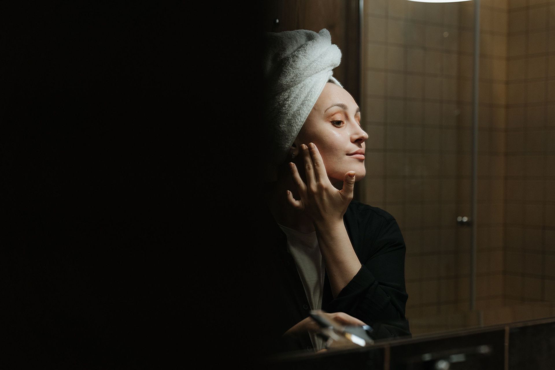 Woman in a black robe applying lotion in front of a mirror, wrapped in a white towel