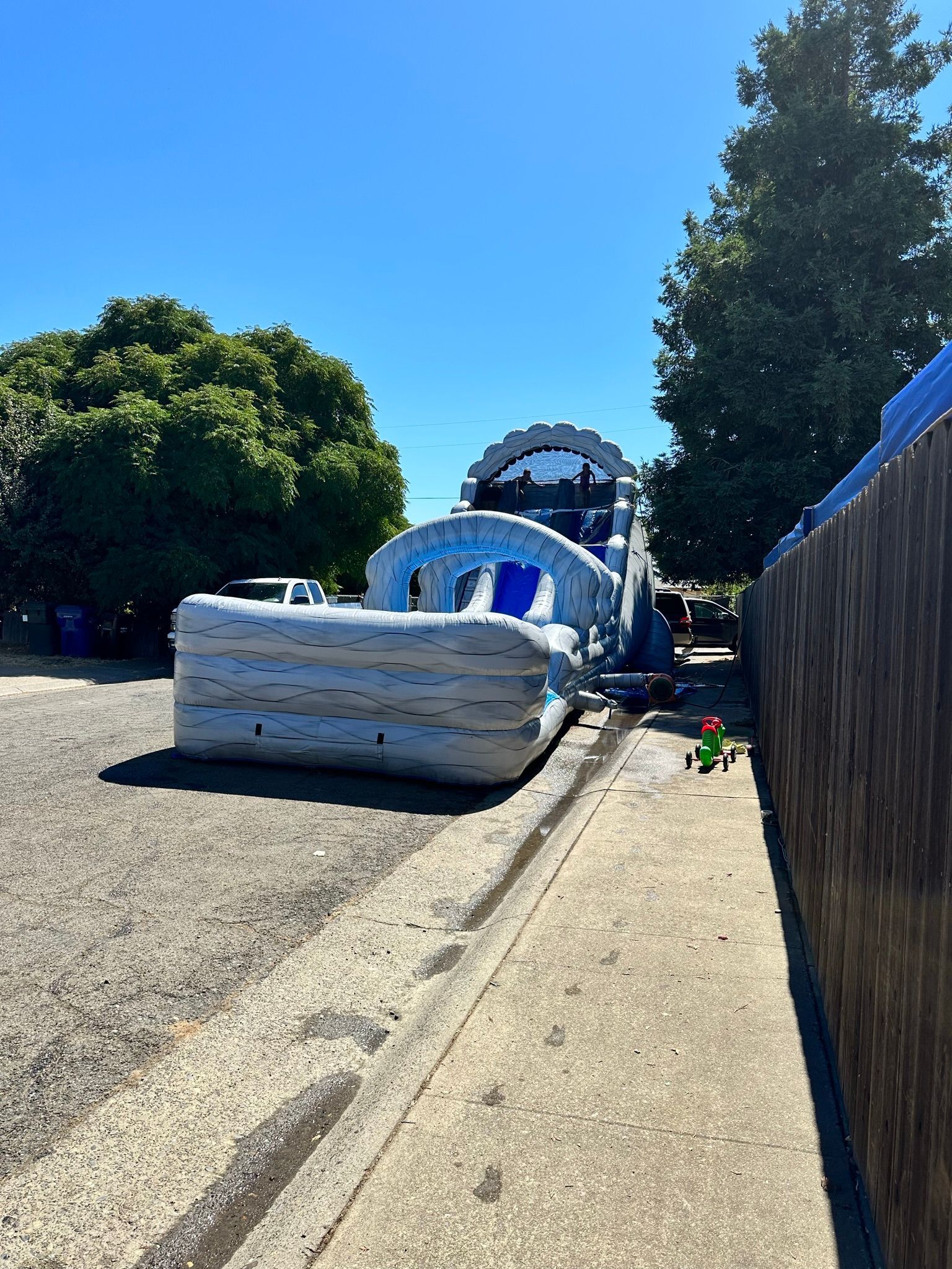 Inflatable bouncy obstacle course on a gravel path beside trees and a wooden fence under a clear blue sky