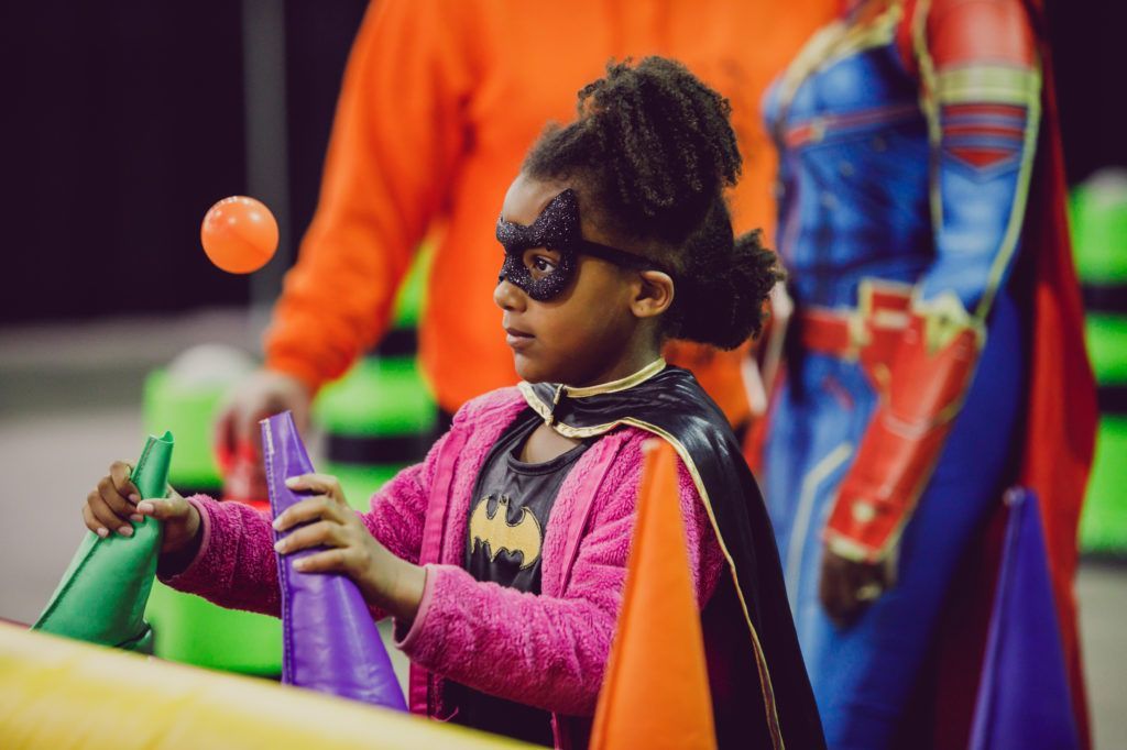 Child wearing sunglasses and a pink jacket stands by colorful inflatable play equipment with a superhero figure behind.